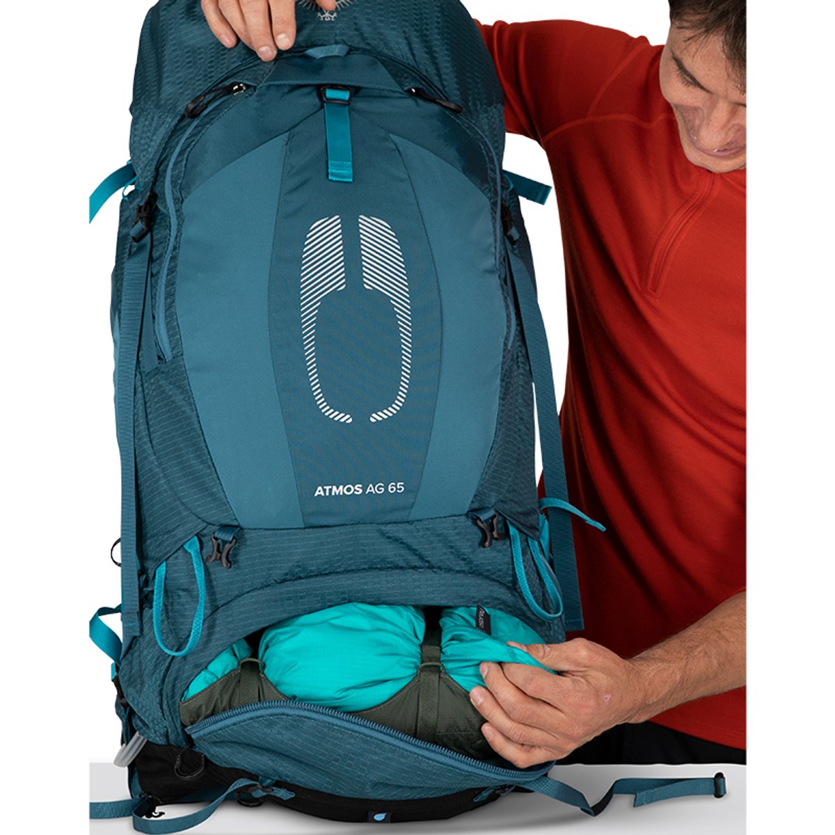 A person in an orange shirt demonstrates the bottom sleeping bag compartment of a blue Osprey Atmos AG 65 pack, showing green gear tucked inside the zippered opening.