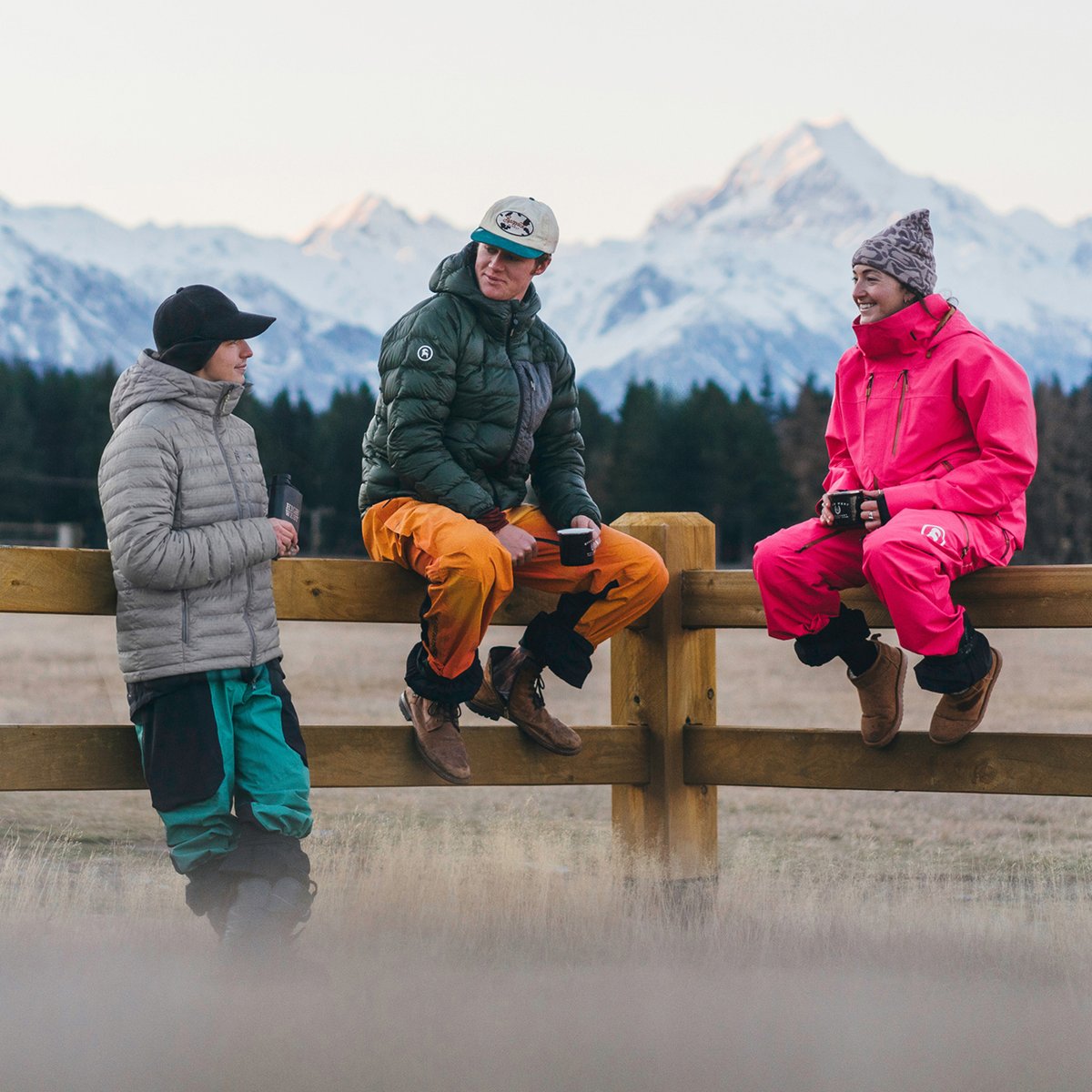 Three friends in insulated jackets and ski pants gather on a wooden fence for après with mugs, snow-covered peaks behind them.