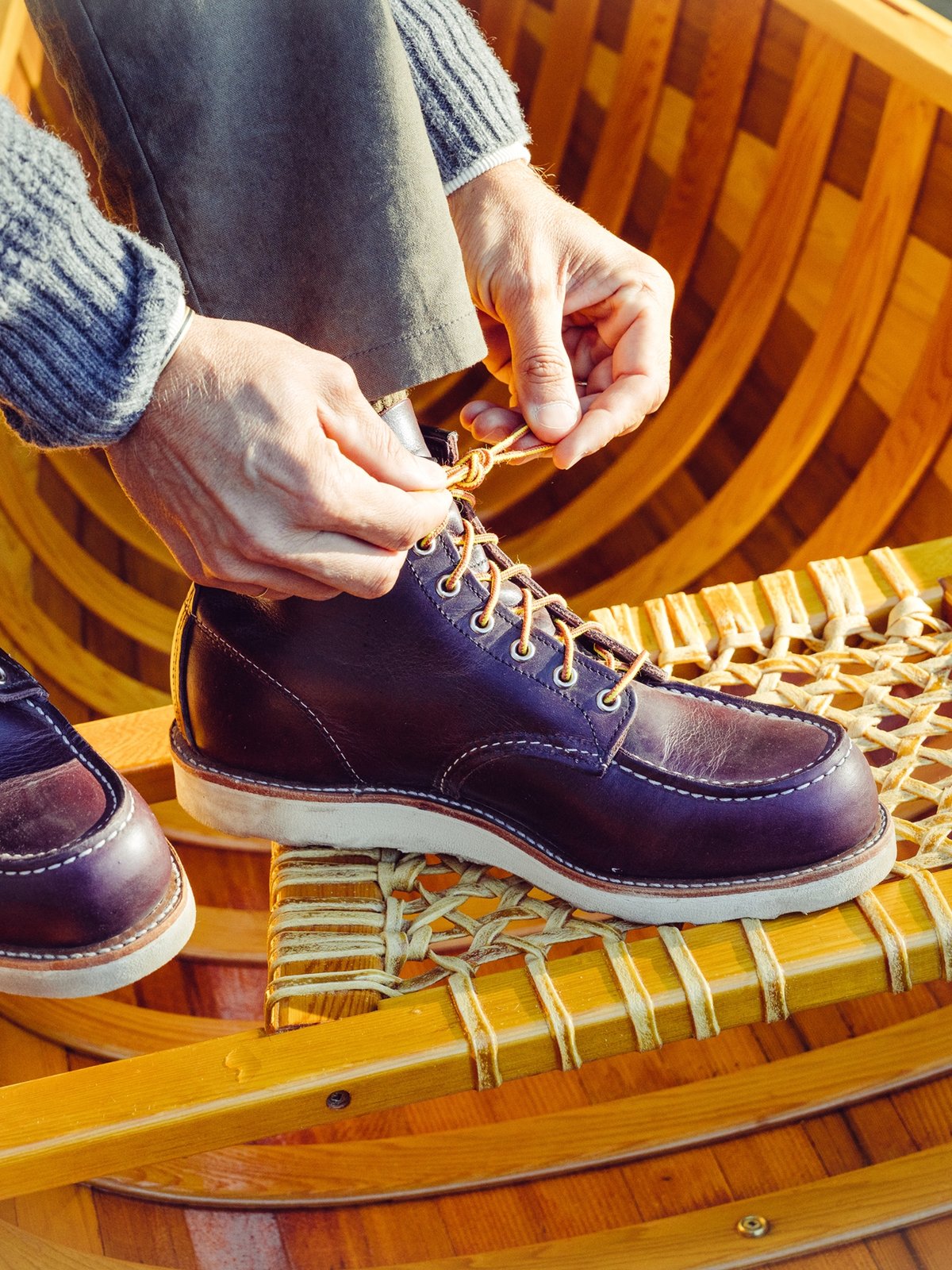 A person wearing olive green pants and a gray sweater is sitting in a wooden boat, tying the laces of a dark brown Redwing leather boot.