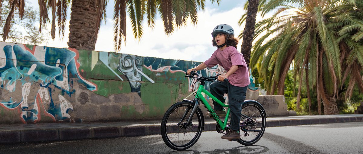 Kid wearing helmet riding down paved road with graffitied wall and trees in the background.