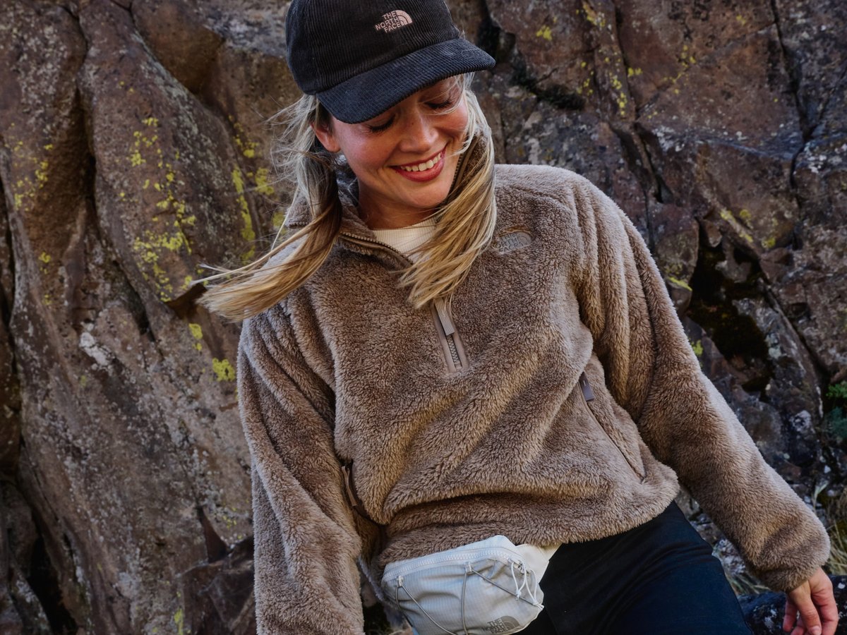 Smiling hiker in a North Face sherpa fleece and corduroy cap leans against lichen-covered granite, fanny pack at the waist.
