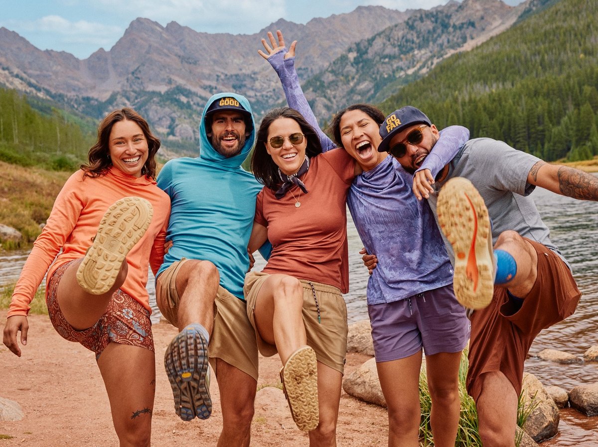 Five friends kick up their heels laughing beside a mountain lake, wearing colorful sun hoodies and trail shoes, peaks rising behind them.