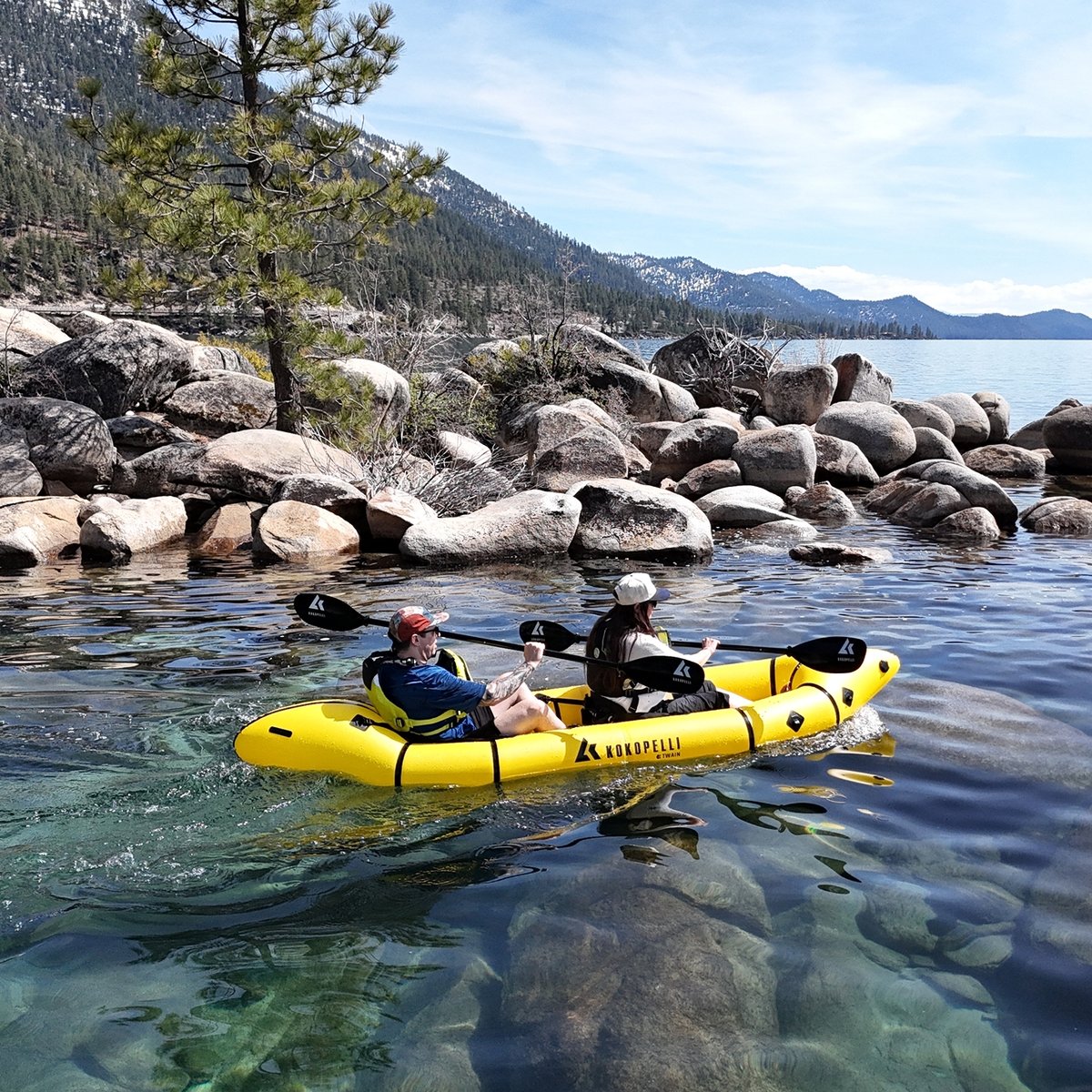 Two people paddle a yellow inflatable kayak on a clear lake near a rocky, tree-lined shore.