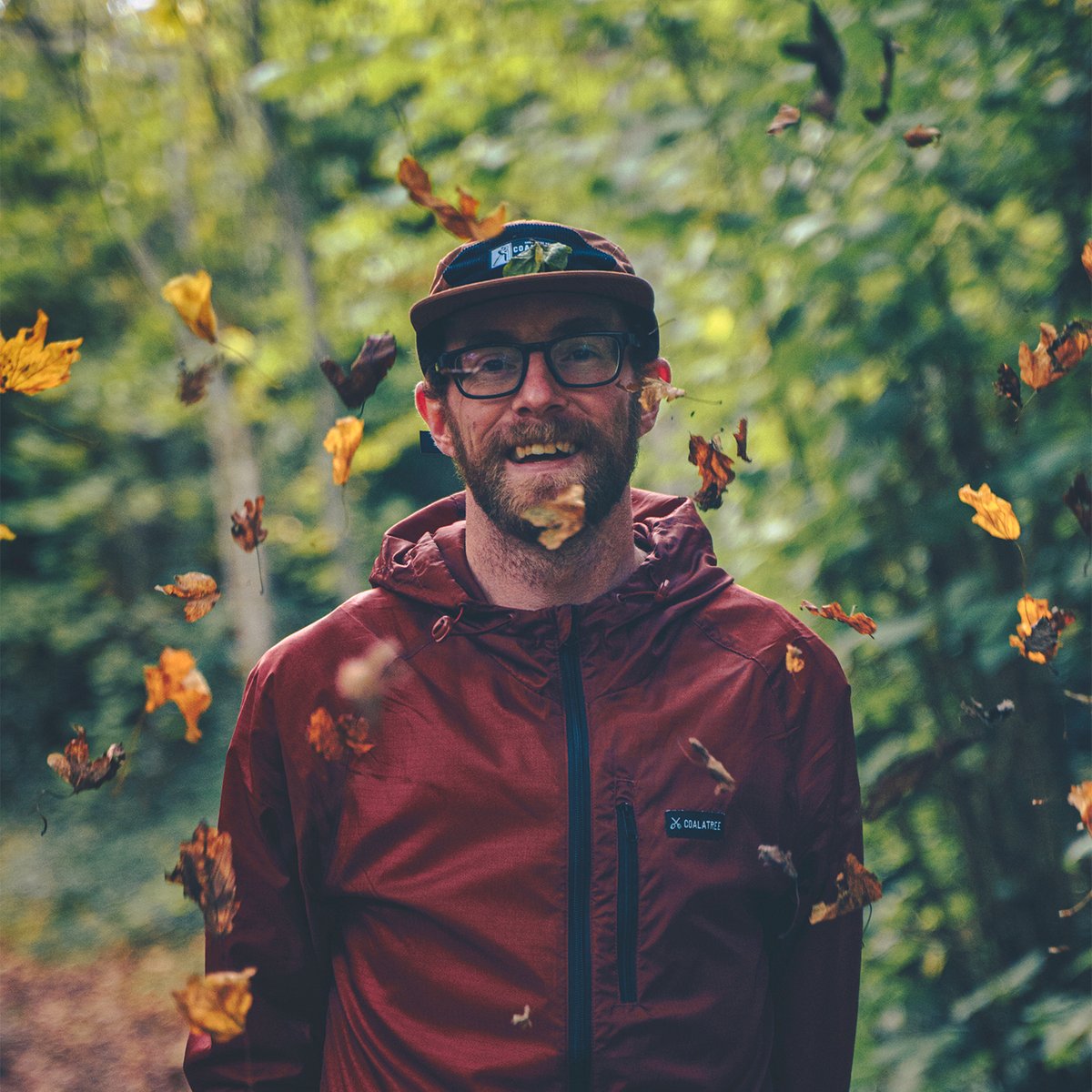 Person smiling wearing Coalatree workwear hat stadning in a forest while yellow leaves fall around them.