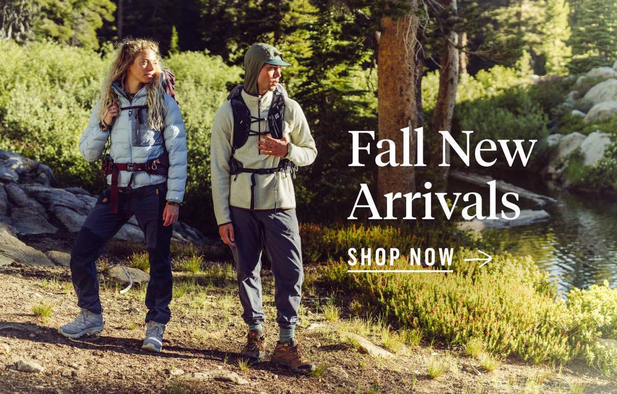 Two people in outdoor hiking gear stand in a forested area near a pond, with text overlaid reading "Fall new arrivals. Shop now."