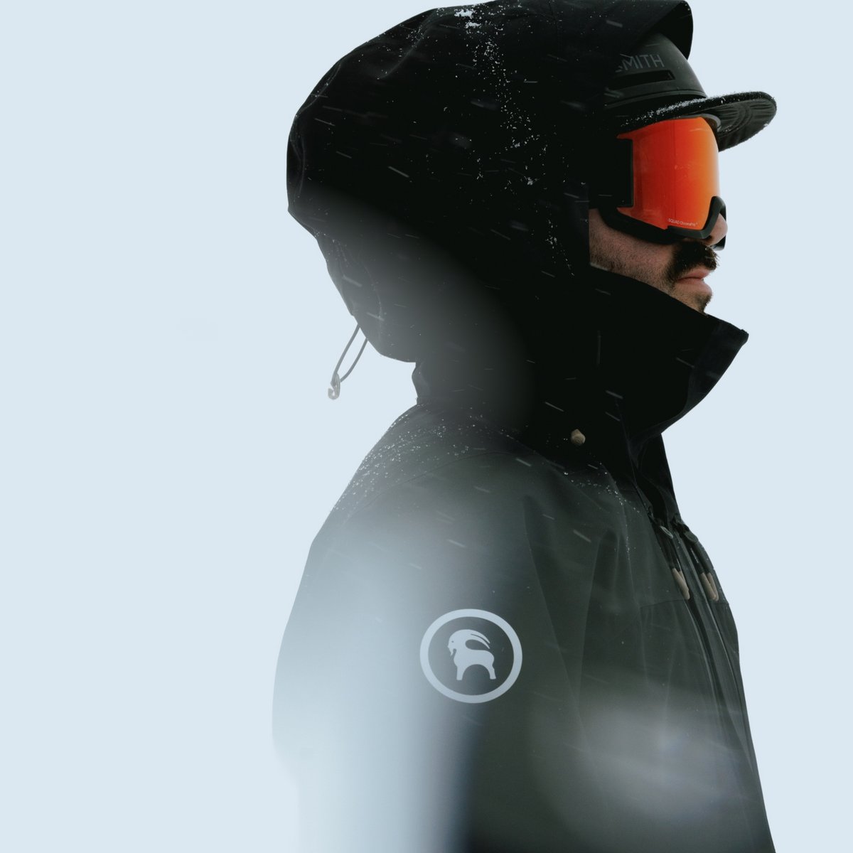 Profile of a skier in a black hooded shell and orange-lens goggles, with snow falling against a pale, overcast sky.