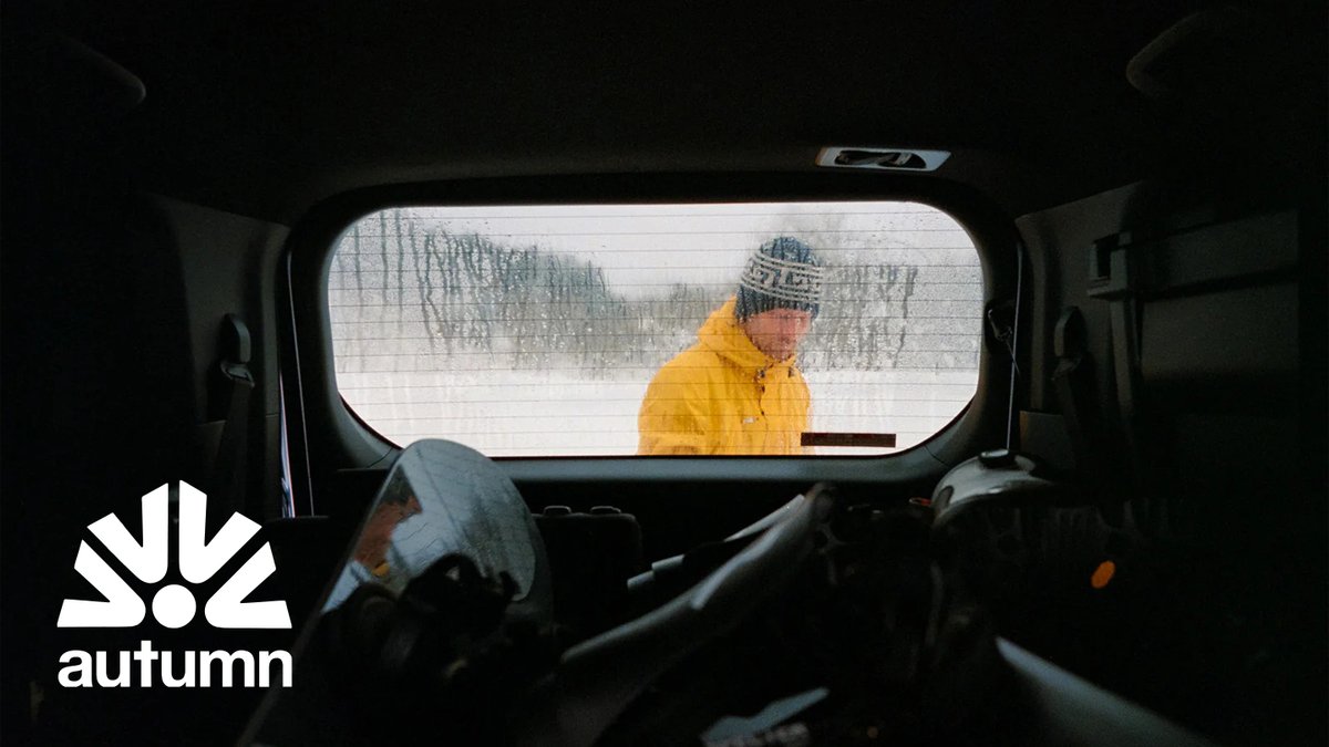 A person wearing a yellow winter jacket and Autumn knit beanie stands in a snowy mountain landscape viewed through a vehicle window, showcasing cold-weather outdoor apparel.
