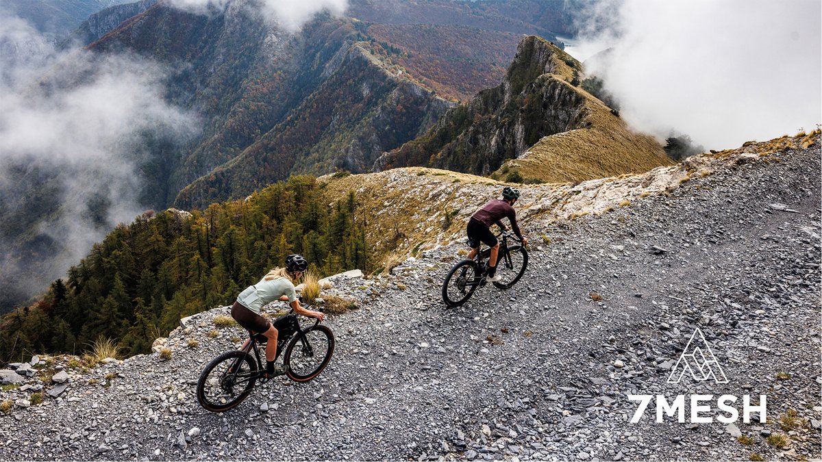 Two cyclists ride gravel bikes on a mountain trail with misty peaks and alpine forest in the background during adventure cycling.