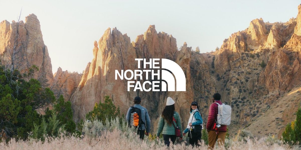 Four people hike through a grassy, rocky canyon, with The North Face logo displayed in the center of the image.