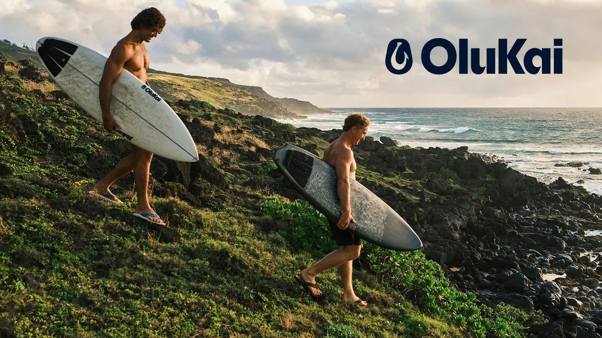 Two shirtless surfers carry their boards down a rocky, vegetation-covered coastline toward the ocean on a sunny day.