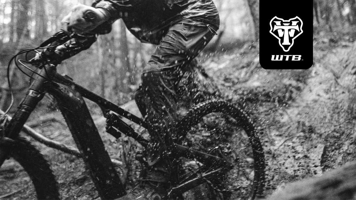Mountain biker performing a muddy trail descent through a forested path wearing protective gear and riding a full-suspension mountain bike.