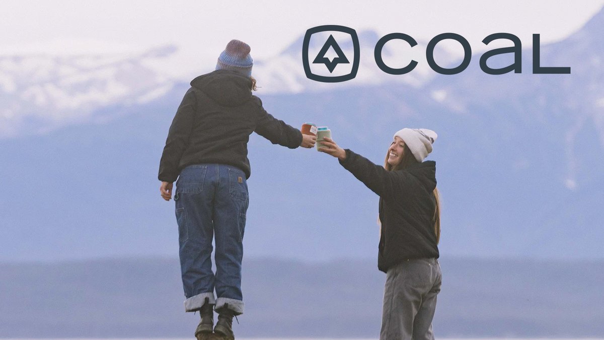 Two people wearing winter Coal Headwear beanies and jackets give each other cheers with their drinks, set against a mountain landscape.
