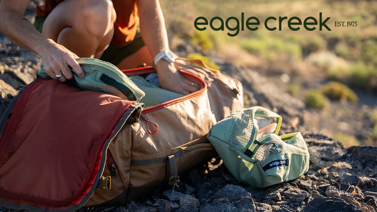 A climber organizes climbing gear including rope and harness from their Eagle Creek backpack while sitting on rocky terrain.