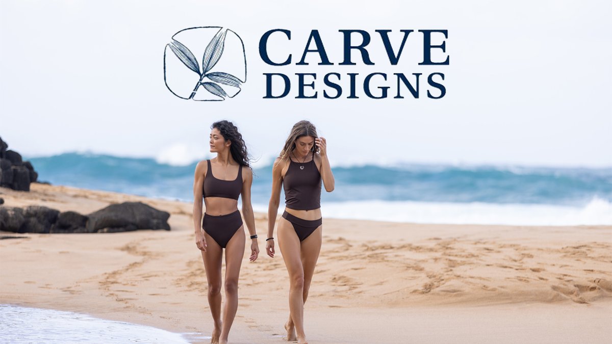 Two women wearing brown swimsuits walk along a sandy beach with the Carve Designs logo centered at the top.