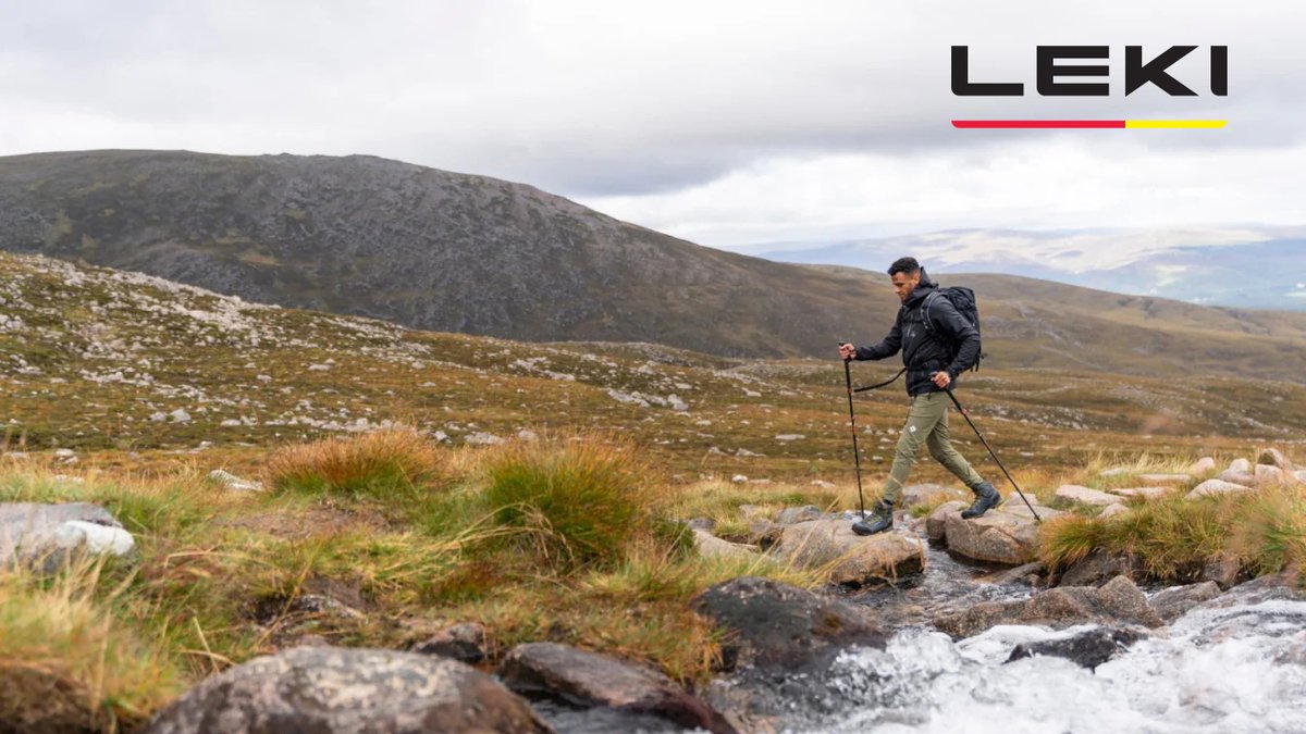 A hiker in black jacket and khaki pants uses trekking poles to navigate across rocky alpine terrain with mountains and sparse vegetation in the background.