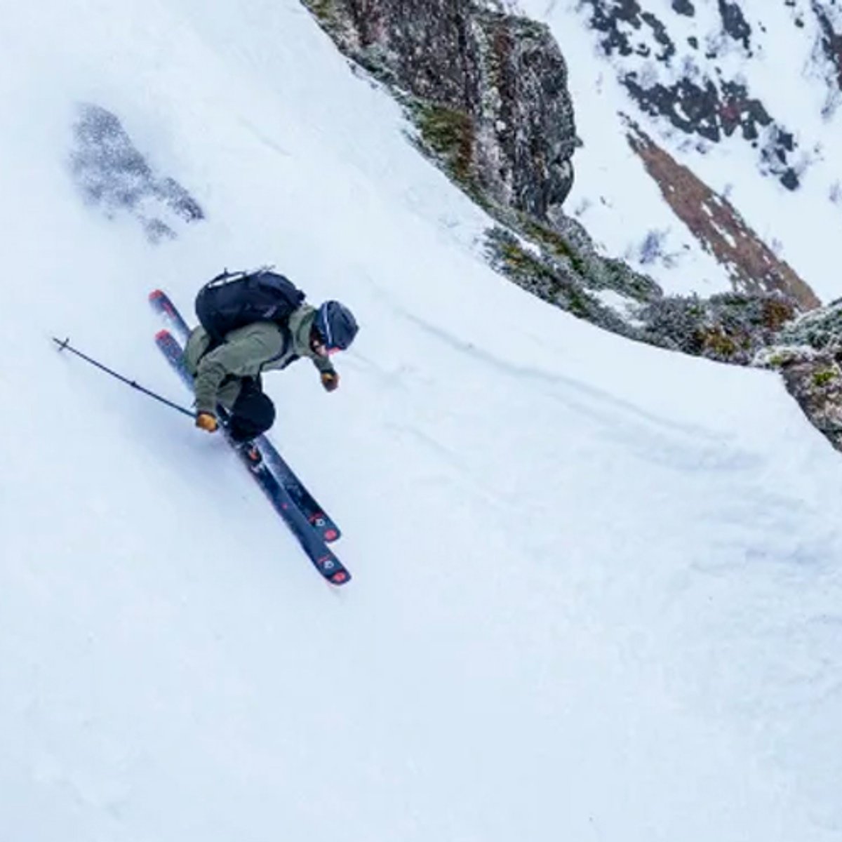 Skier descending a snowy mountain slope with rocky terrain and alpine peaks in an overhead aerial view.