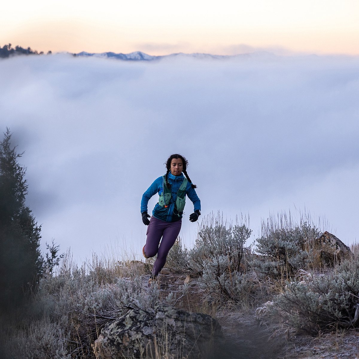 A person trail-runs on a frosty mountainside, high above a valley filled with clouds.