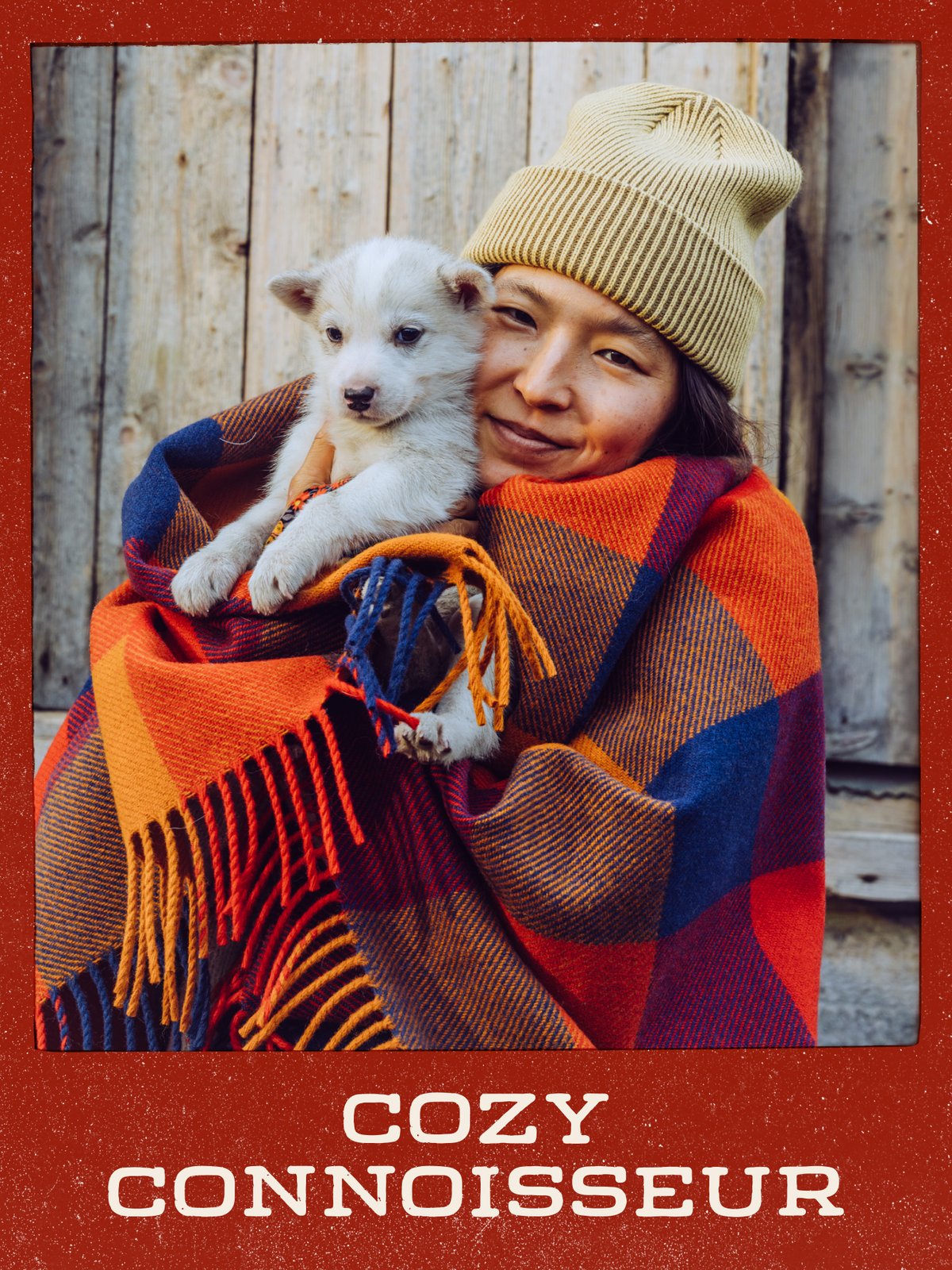 Person in a beanie wrapped in a plaid wool blanket, holding a white puppy against a weathered wood wall: “Cozy Connoisseur.”