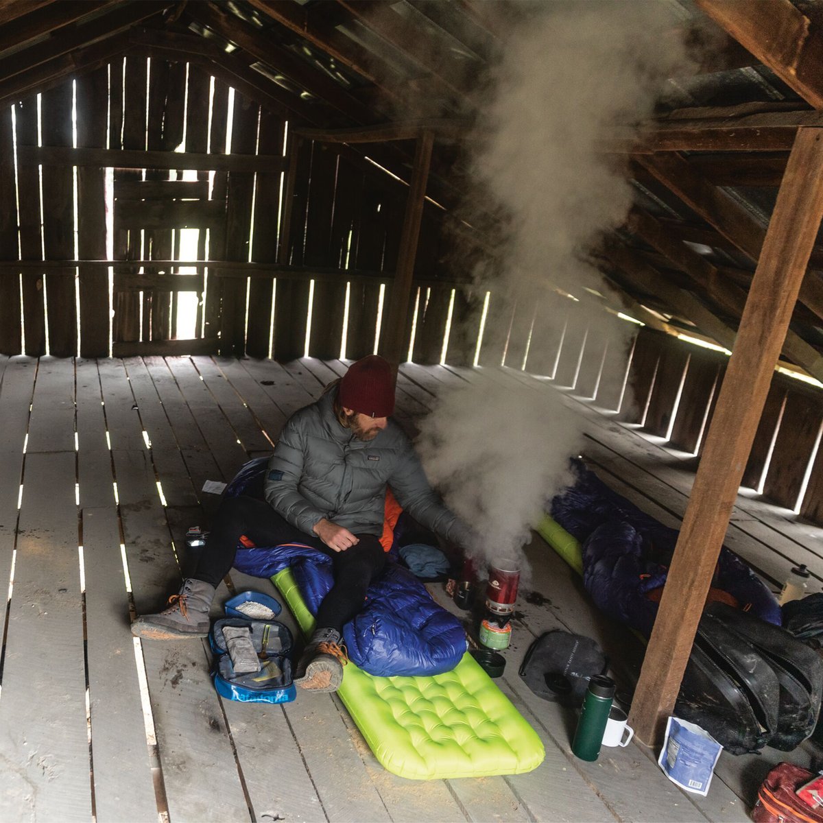 A hiker in winter gear sits inside a rustic wooden shelter, cooking with a portable camping stove surrounded by sleeping bags and outdoor equipment.