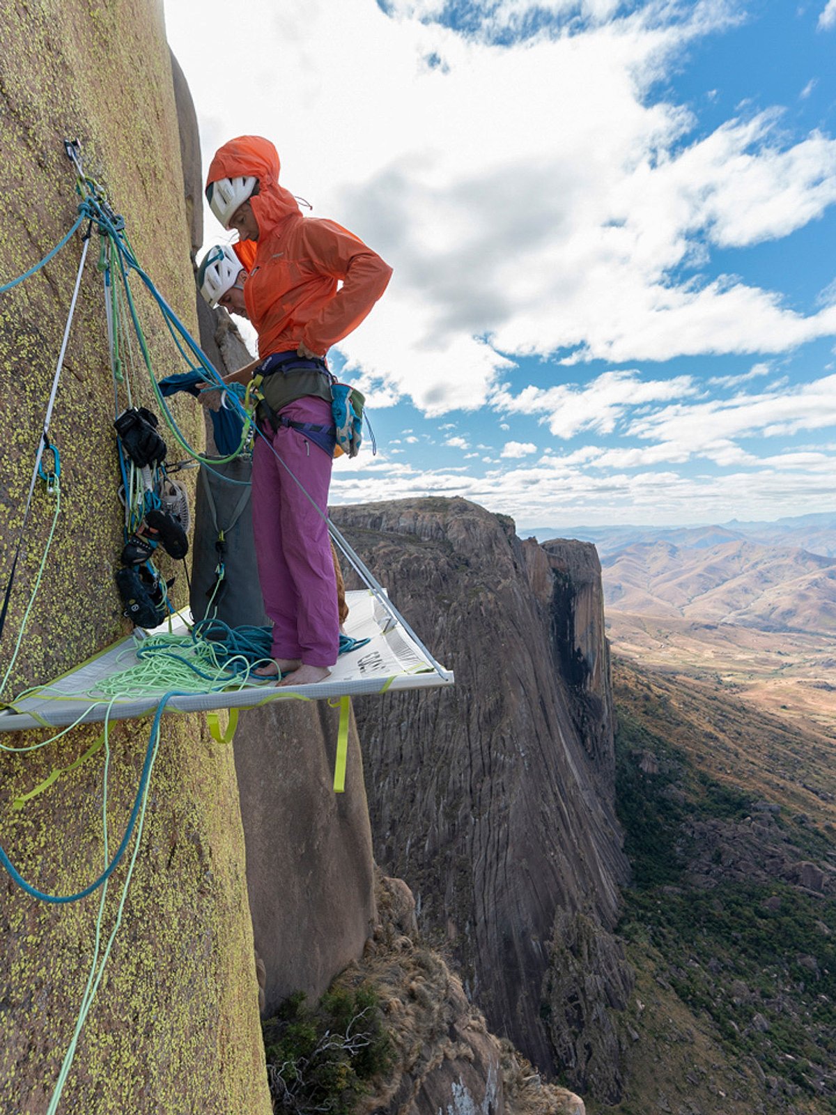 Big wall climber standing on portaledge wearing hooded shell and harness on vertical granite face in windy alpine conditions.