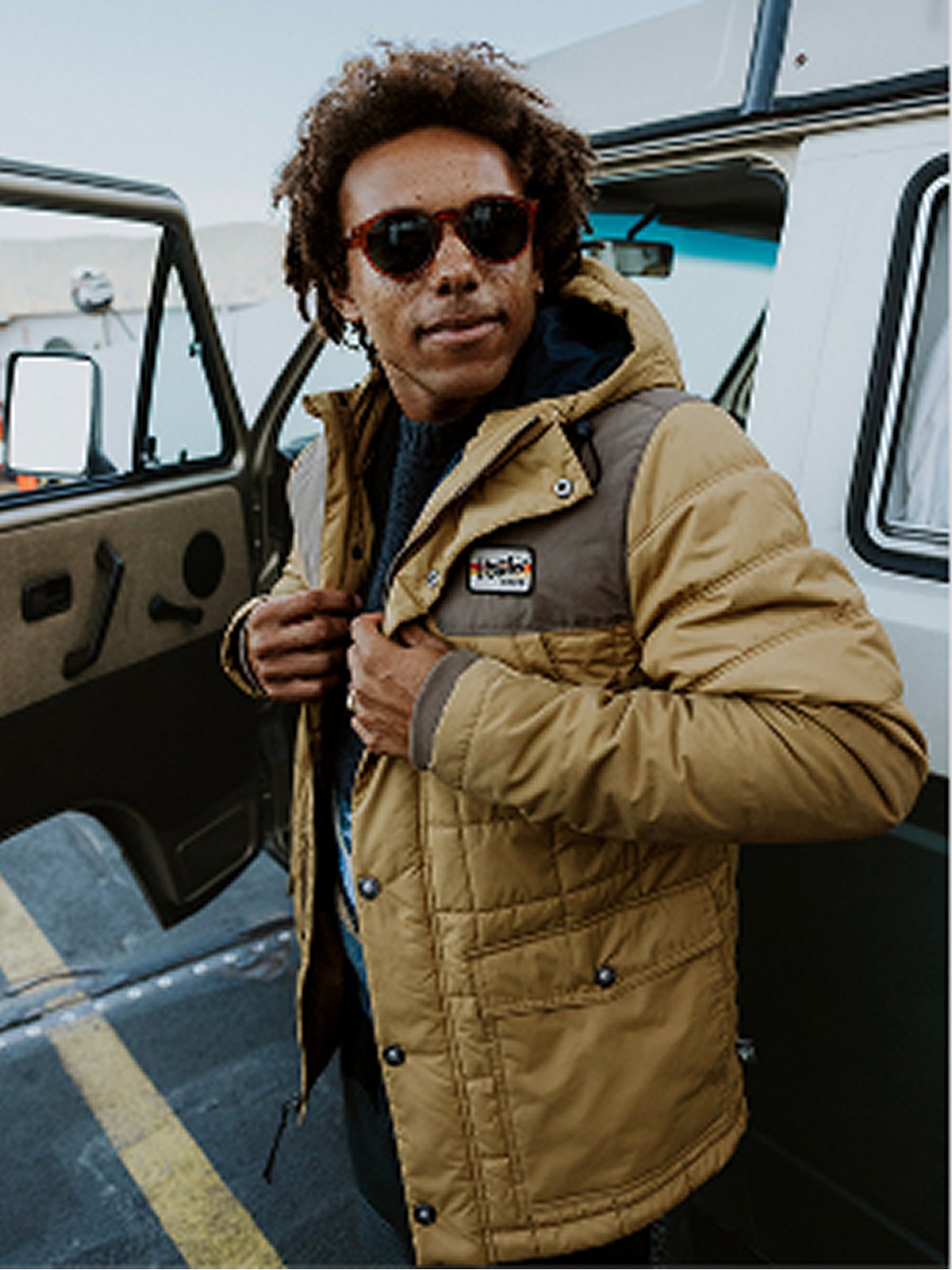 A man wearing a yellow quilted jacket over a light gray hoodie in a pine forest.