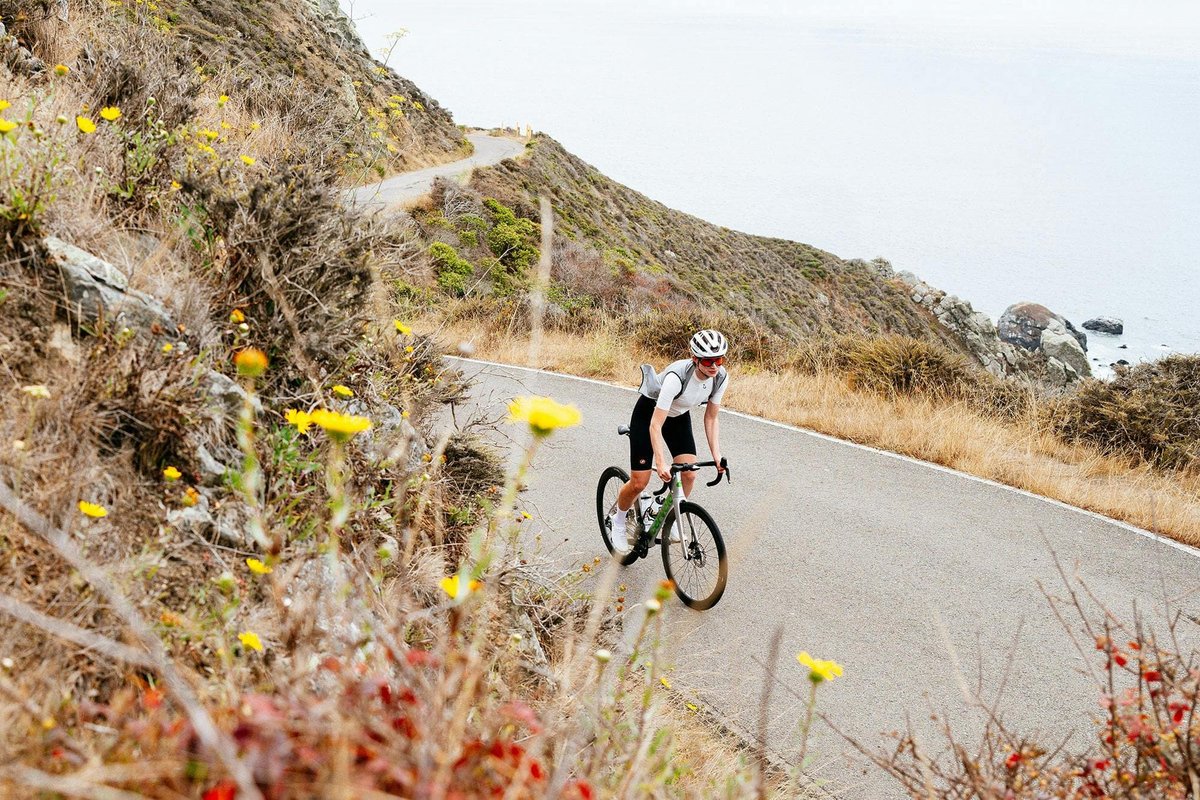 Road cyclist wearing cycling jersey, bib shorts, and road helmet climbing narrow coastal road above rocky ocean shoreline