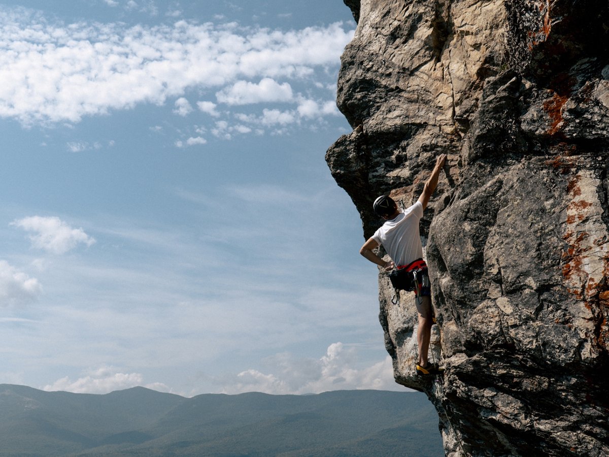 Climber ascending a rocky cliff face with mountain and sky background