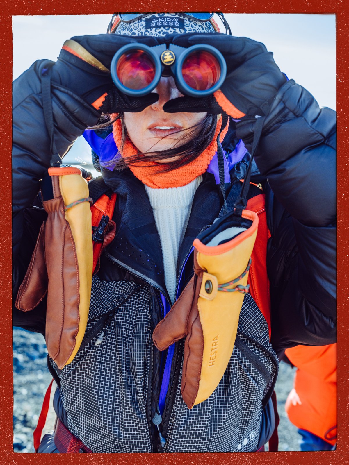 Person in a puffy jacket looking through binoculars, with leather mittens hanging at their chest.