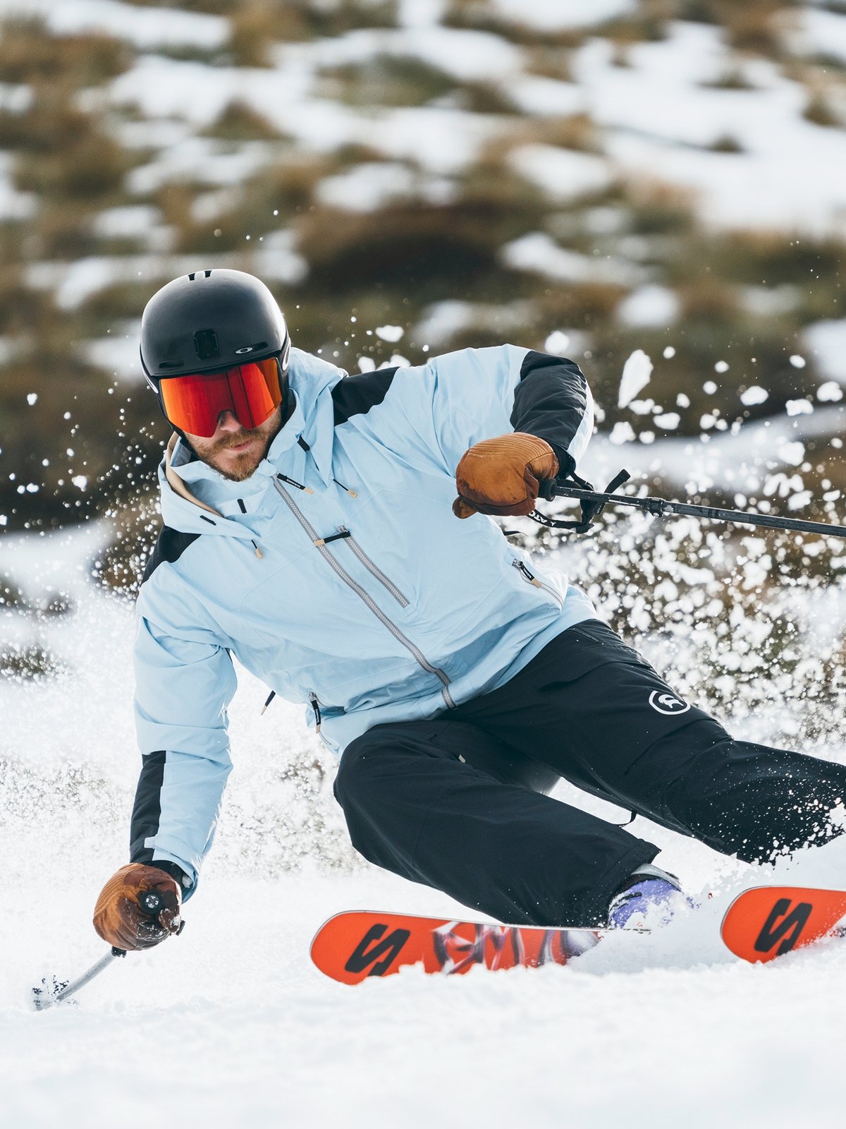 A skier wearing a light blue jacket, black pants, black helmet, and red goggles carves sharply, creating a spray of snow with orange skis.
