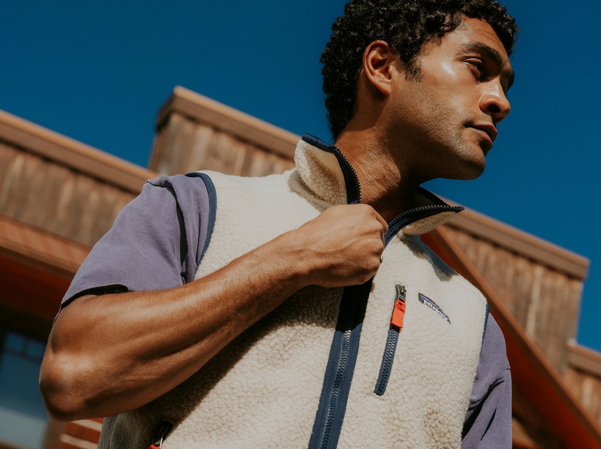 A person wearing a light fleece vest with a zippered chest pocket over a short-sleeved shirt stands outdoors in front of a wooden building, holding the zipper of the vest.