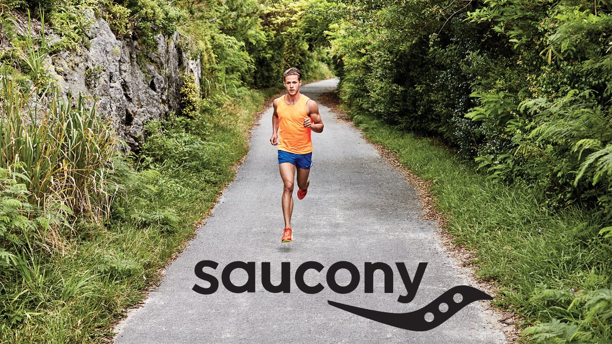 Male runner in orange shirt and blue shorts jogging on a scenic forest trail lined with green vegetation and stone walls while wearing Saucony running shoes.