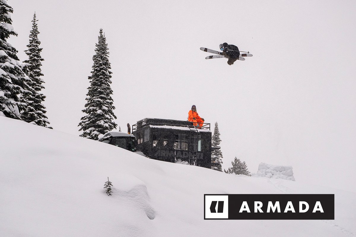 One skiier jumps high in the air, while the other watches while sitting on top of a truck.