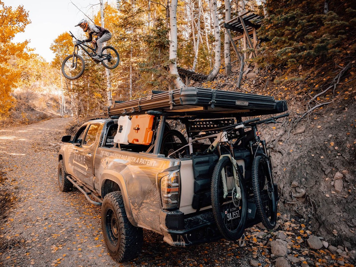 Tan Tacoma with rooftop tent and two bikes on a CACHE tailgate pad, rider jumping a wooden feature in fall aspens.