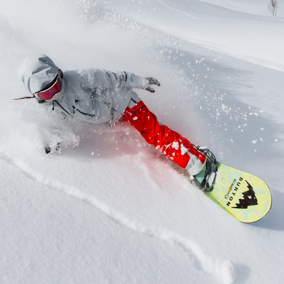 A snowboarder in a gray jacket and red pants rides through powder snow, with snow spraying around the yellow Burton snowboard.