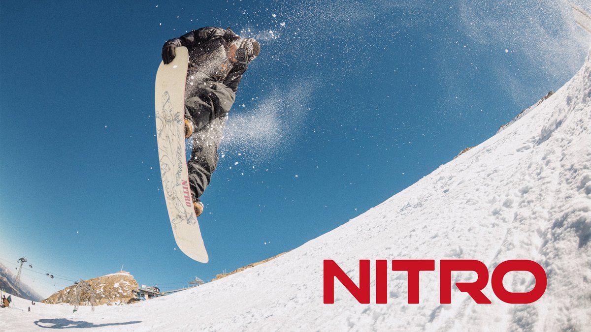 Nitro snowboarder launching high into the air off a snowy mountain slope with a white board against a clear blue sky, showcasing extreme winter sports action and NITRO gear performance.