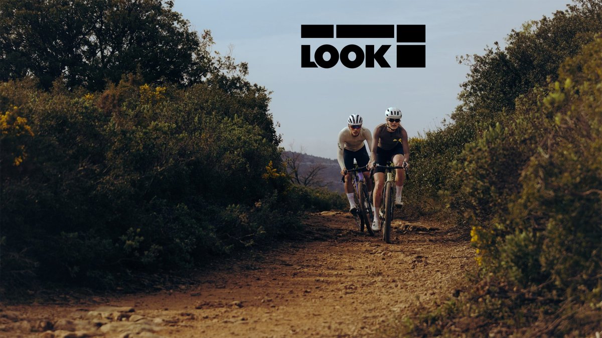 Two cyclists ride side-by-side on a scenic dirt trail through golden shrubland wearing helmets and Look Cycle athletic gear during daytime.