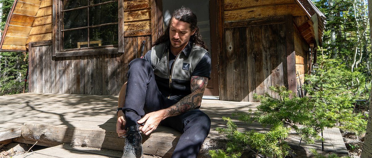 Person wearing Coalatree 10-Gauge Vest sitting on cabin porch in teh forest adjusting his boot. 