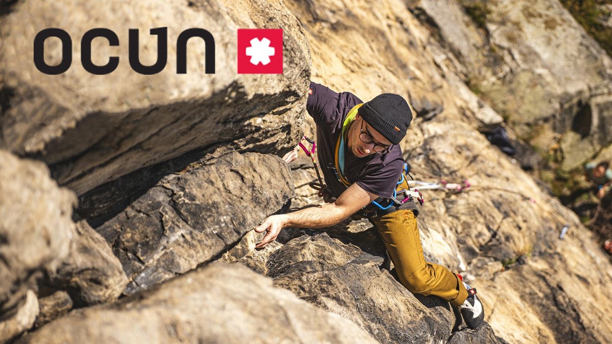 A male rock climber wearing OCUN gear scales a steep outdoor rock face while secured with safety equipment and carabiners.