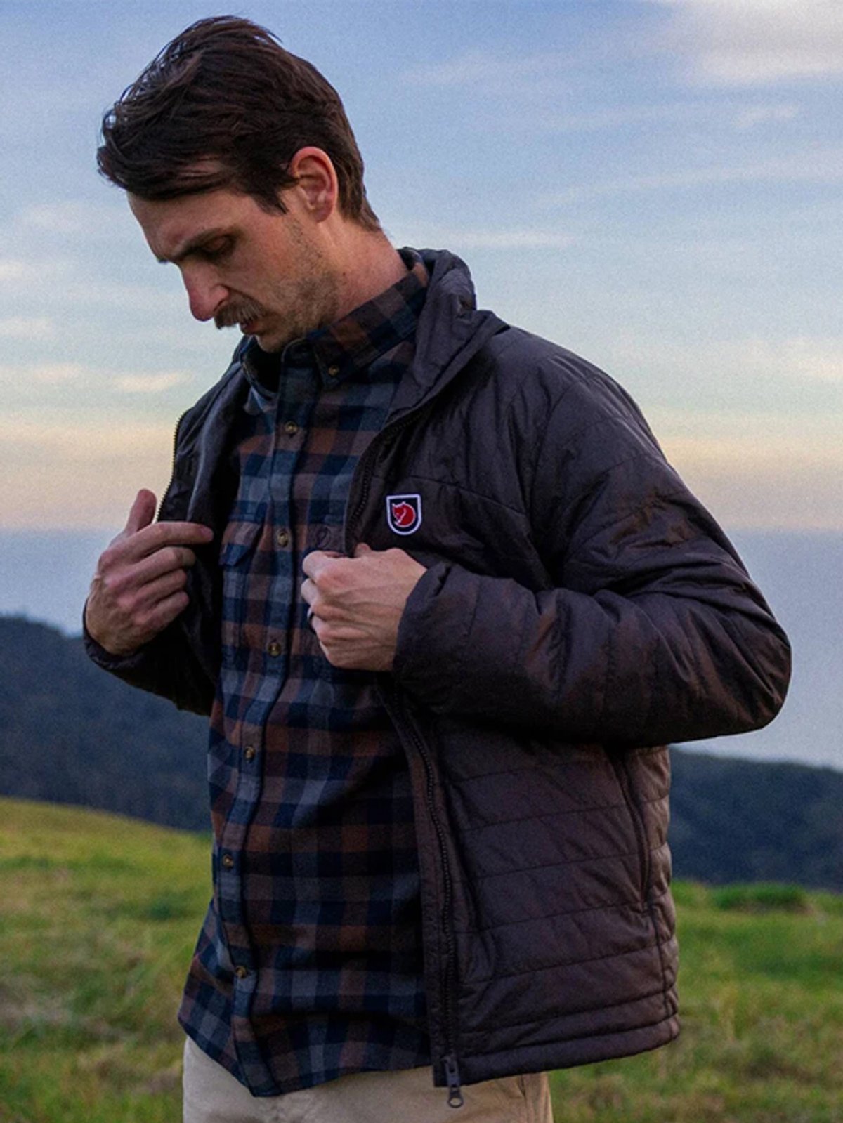 A man wears the Fjallraven X-Latt Jacket against a mountain background.
