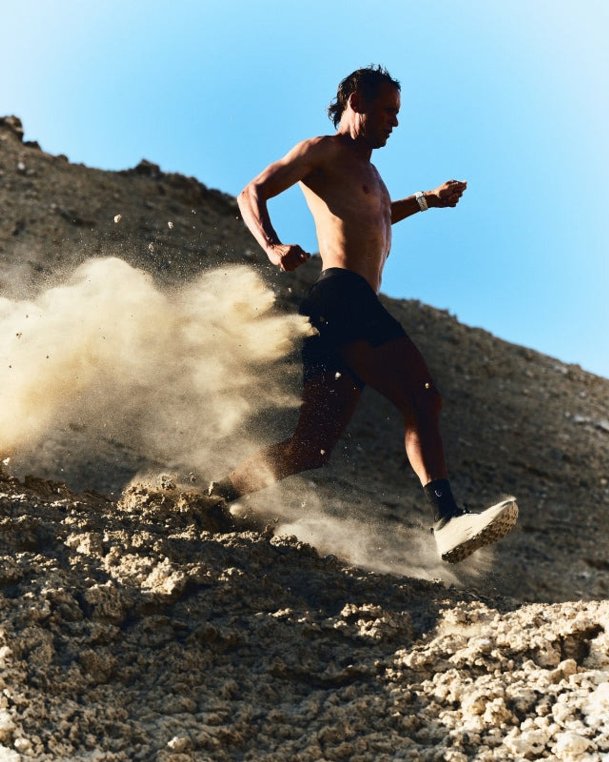 Shirtless male runner sprinting across a crumbling dirt slope with a large dust cloud billowing behind, against a blue sky.