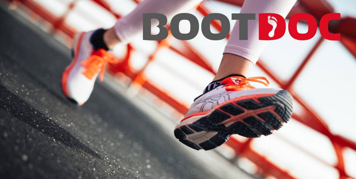 Runner wearing red and orange athletic shoes mid-stride on an asphalt path with red and white training cones in the background.