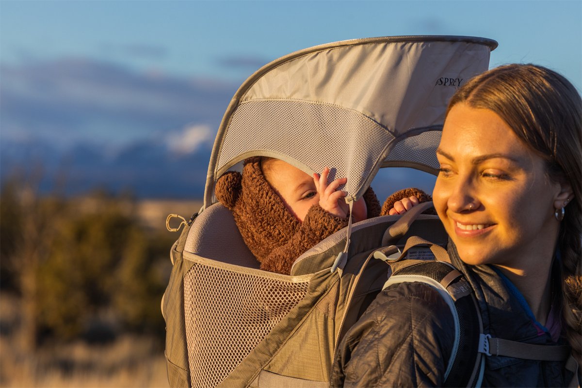 Gemini said A woman smiles while carrying a baby in an Osprey hiking carrier during a sunset. T