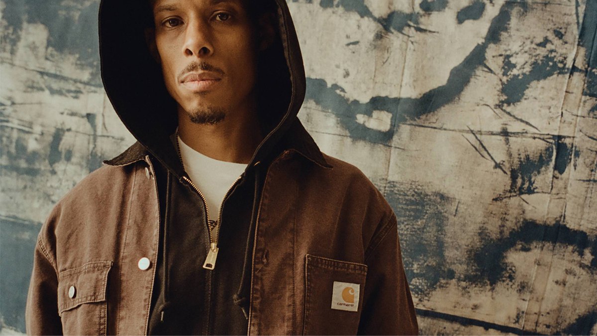A man wears a dark brown Carhartt hooded work jacket with the brand's signature logo on the pocket.