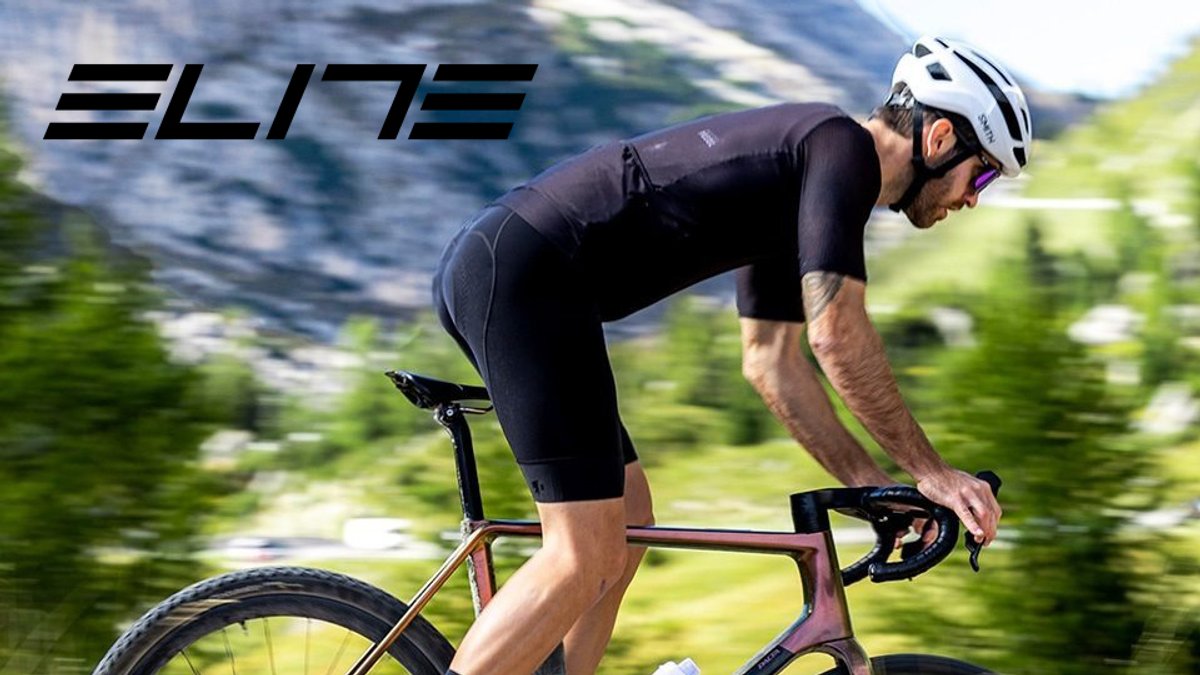 Male cyclist in black Elite sportswear riding a purple road bike through mountainous terrain while wearing a white helmet.