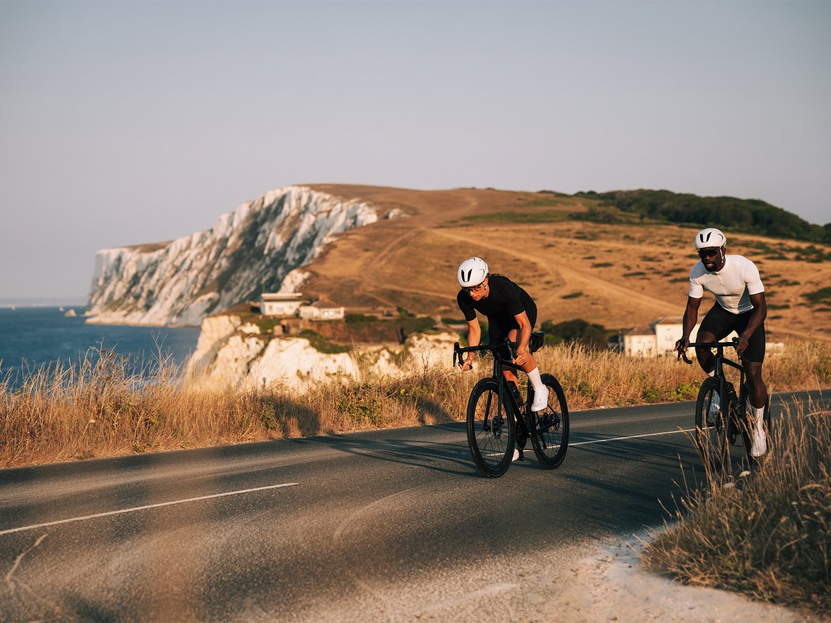 Two cyclists riding side by side along a coastal road, ocean horizon in the background, white S-PHYRE shoes visible on both riders