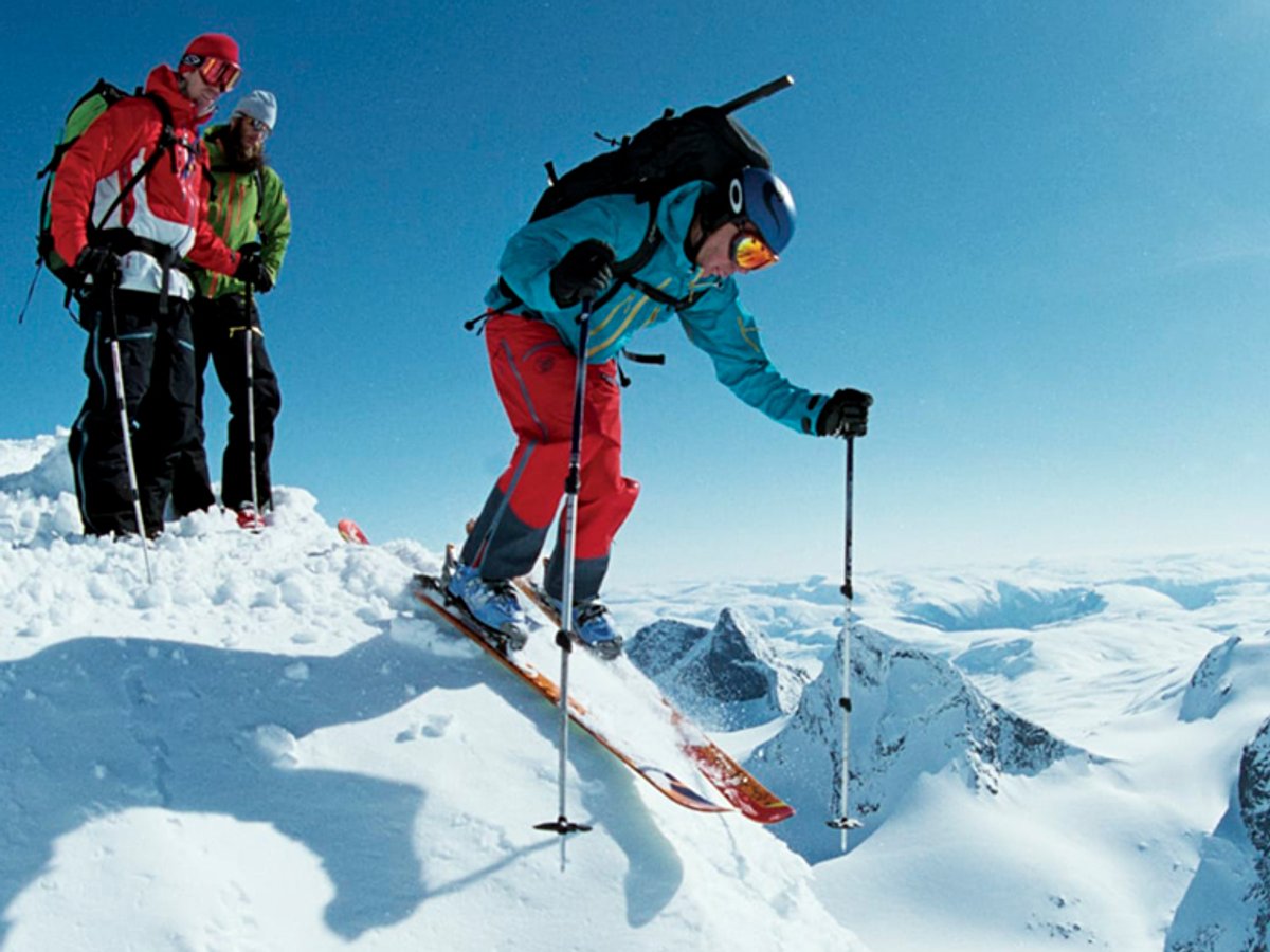 A skier in turquoise and red gear descends a snowy mountain slope while two mountaineers in bright outfits stand on the summit above under clear blue skies.