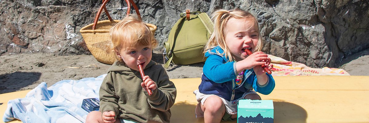 Two toddlers enjoy snacks on a sandy beach with Patagonia gear and provisions nearby on a sunny day.