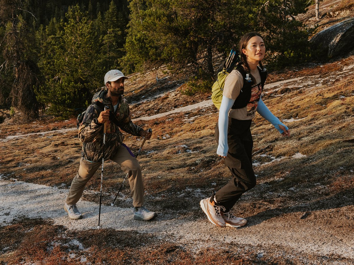Two hikers ascending a rocky alpine trail at golden hour, wearing packs, trekking poles, and layered hiking apparel.