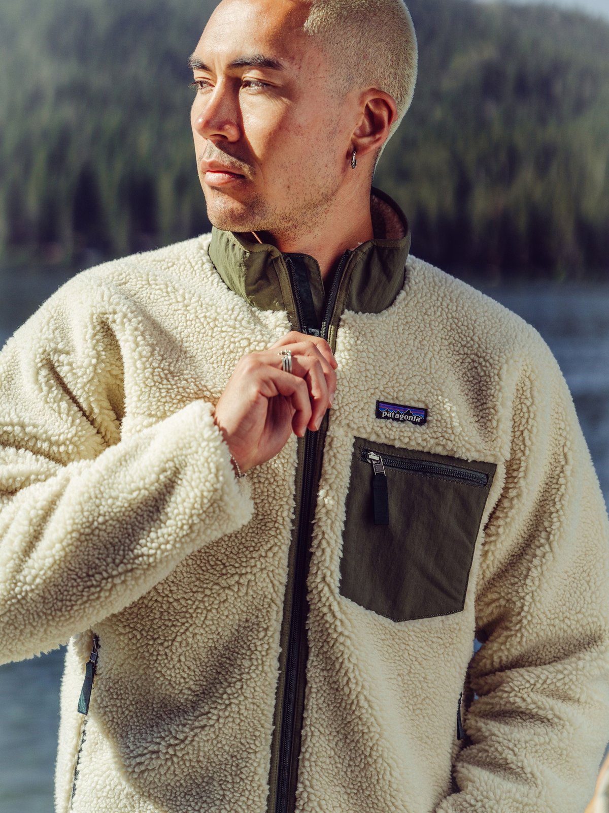 A person stands near a lake wearing a cream-colored sherpa fleece jacket with an olive green chest pocket and collar; the background features forested hills.