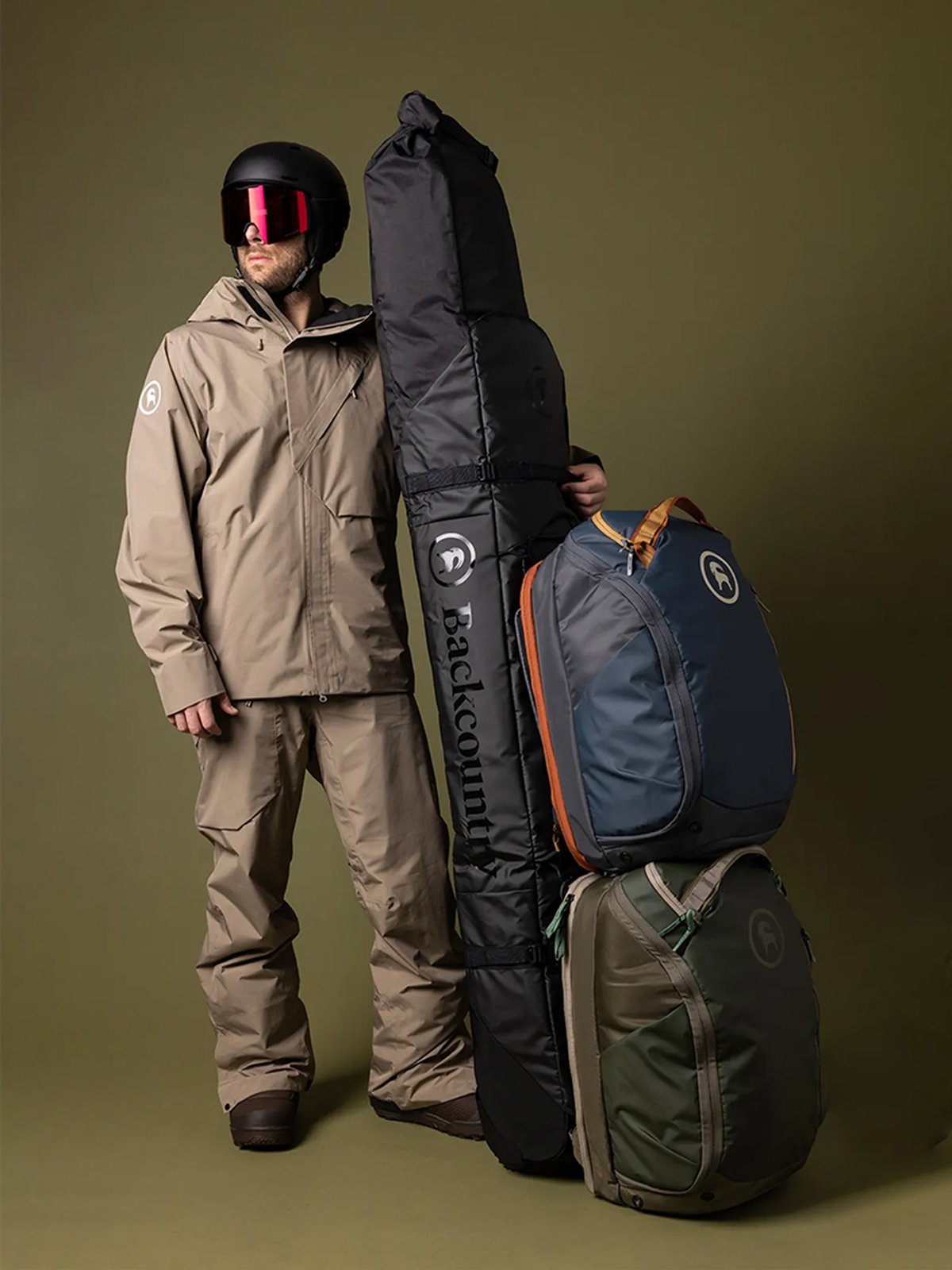 Person in snowabord outerwear wearing helmet and goggles holding rolling ski bag with two boot bags attached.